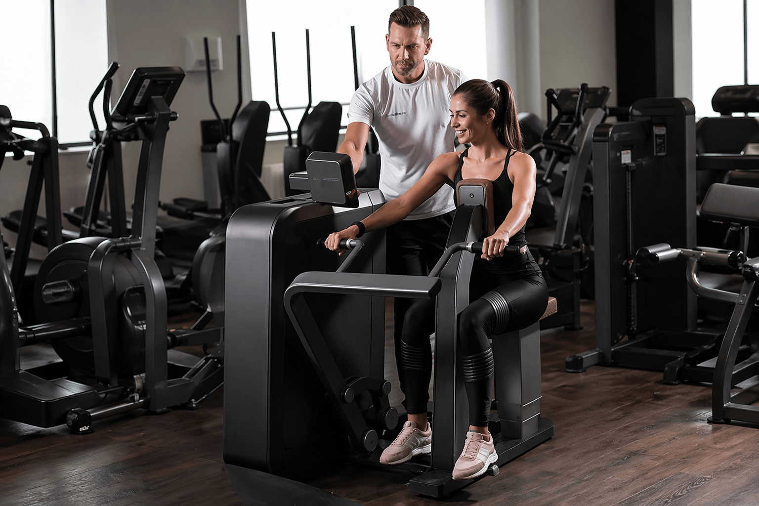 Eine Frau trainiert an einem Rudergerät im Fitnessstudio, daneben steht ihr männlicher Trainer und gibt ihr Hinweise