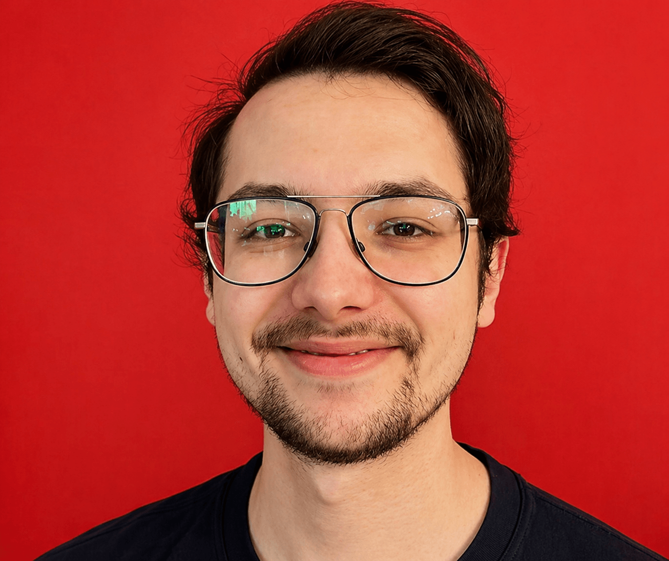 Ein Mann mit Brille und kurzen dunklen Haaren lächelt vor einem leuchtend roten Hintergrund Ein Mann mit Brille und kurzen dunklen Haaren lächelt vor einem leuchtend roten Hintergrund