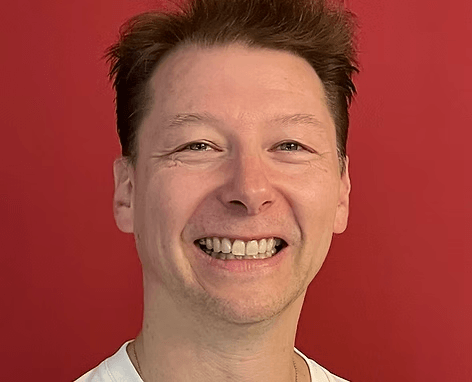 Ein lächelnder Mann mit kurzen braunen Haaren und weißem Hemd steht vor einem einfarbig roten Hintergrund Ein lächelnder Mann mit kurzen braunen Haaren und weißem Hemd steht vor einem einfarbig roten Hintergrund