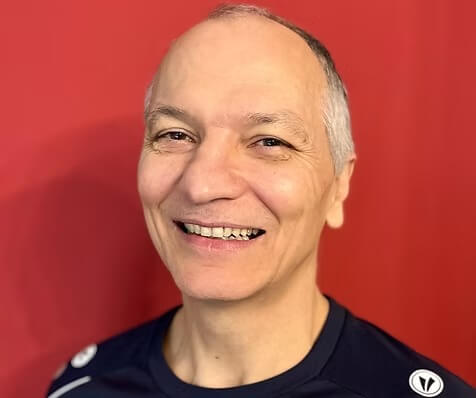 Ein lächelnder älterer Mann mit kurzen grauen Haaren trägt ein dunkles Hemd und steht vor einem einfarbig roten Hintergrund Ein lächelnder älterer Mann mit kurzen grauen Haaren trägt ein dunkles Hemd und steht vor einem einfarbig roten Hintergrund