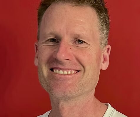 Ein lächelnder Mann mit kurzen hellbraunen Haaren steht vor einem leuchtend roten Hintergrund und trägt ein weißes Hemd Ein lächelnder Mann mit kurzen hellbraunen Haaren steht vor einem leuchtend roten Hintergrund und trägt ein weißes Hemd