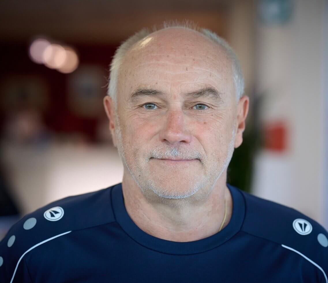 Ein älterer Mann mit kurzem grauem Haar und gestutztem Bart lächelt in einem marineblauen Sportshirt freundlich in die Kamera drinnen