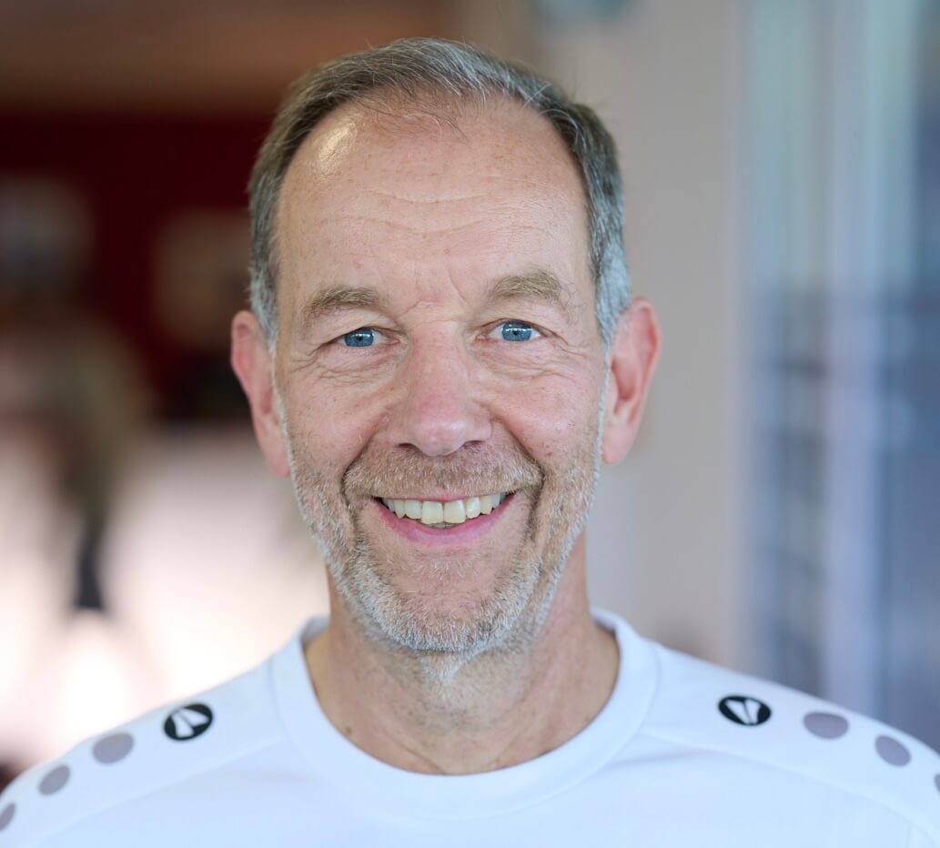 Ein älterer Mann mit grazen Haaren und Bart lächelt in weißem Hemd, steht drinnen, der Hintergrund ist unscharf