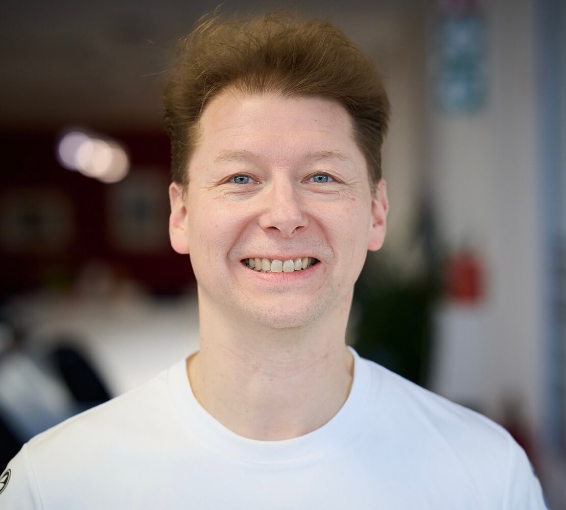 Ein lächelnder Mann mit heller Haut und blauen Augen trägt ein weißes Hemd und steht drinnen vor unscharfem Hintergrund