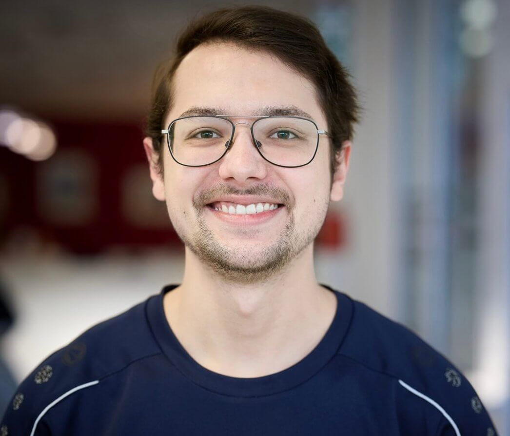Ein junger Mann mit Brille und kurzen braunen Haaren lächelt in die Kamera und trägt drinnen ein marineblaues Hemd