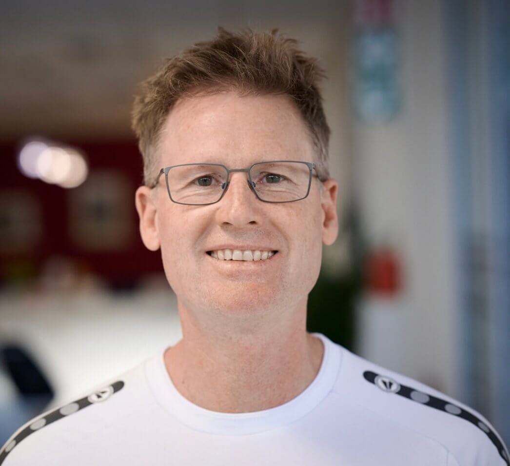 Ein lächelnder Mann mit kurzen hellbraunen Haaren und Brille trägt ein weißes Hemd und steht drinnen vor unscharfem Hintergrund
