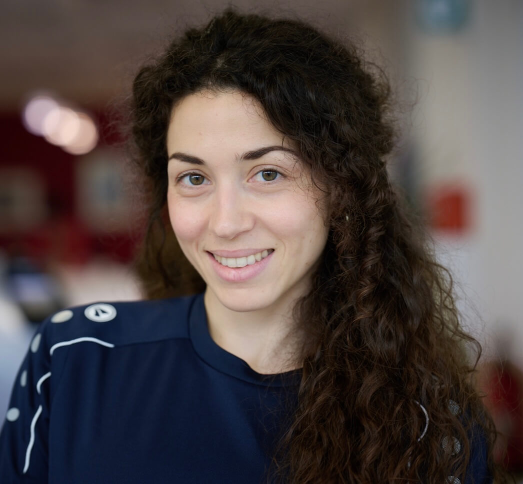 Eine Frau mit langen lockigen Haaren und dunkelblauem Shirt lächelt in die Kamera, im Hintergrund ein weich verschwommener Innenraum