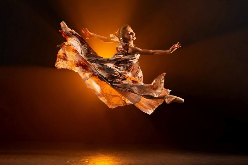 Eine Tänzerin in einem im Wind wehenden Kleid springt elegant durch die Luft, warmes, orange-gelbes Bühnenlicht beleuchtet sie